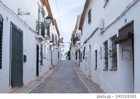 Charming cobblestone street winding uphill through Altea old town with traditional white houses and decorative streetlights under a bright sky, perfect for a Spanish holiday exploration Charming cobblestone street winding uphill through Altea old town with traditional white houses and decorative streetlights under a bright sky, perfect for a Spanish holiday exploration 143528395