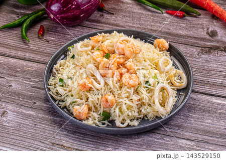 Seafood fried rice with shrimp and squid rings on dark plate on rustic wooden table with eggplant, okra and chili peppers, Asian cuisine dish 143529150