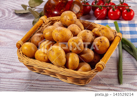 Woven basket filled with unpeeled golden potatoes on weathered wooden table beside cherry tomatoes and green onions 143529214