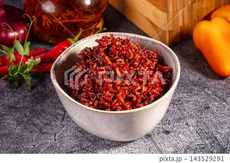 Rustic dark tabletop setting ceramic bowl filled with cooked red rice grains alongside chili peppers and yellow bell pepper as wholesome grain meal 143529291