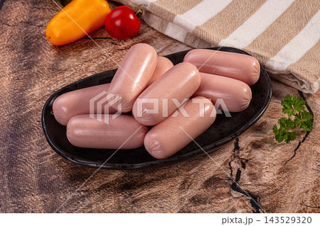 Raw mini frankfurter sausages piled on black oval plate with striped kitchen towel, yellow pepper, tomato and parsley on rustic stone background, food still life 143529320