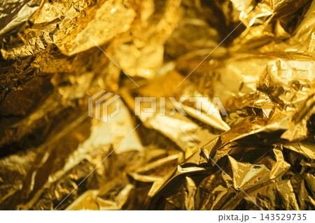 Abstract close up of crumpled reflective gold foil with angular folds, metallic highlights, deep shadows, and richly textured surface filling the entire frame Abstract close up of crumpled reflective gold foil with angular folds, metallic highlights, deep shadows, and richly textured surface filling the entire frame 143529735
