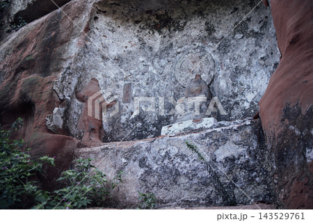 Ancient stone carvings of a seated Buddha and a figure on a rock face surrounded by greenery in a serene outdoor setting Ancient stone carvings of a seated Buddha and a figure on a rock face surrounded by greenery in a serene outdoor setting 143529761