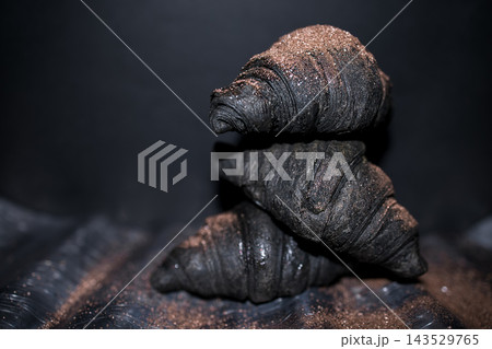 Stacked black croissants with a shiny surface and a dusting of fine brown sugar on a dark background, showcasing unique pastry presentation and texture Stacked black croissants with a shiny surface and a dusting of fine brown sugar on a dark background, showcasing unique pastry presentation and texture 143529765