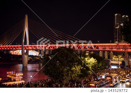 Night view of a modern suspension bridge illuminated in red, with a bustling waterfront and city skyline featuring tall buildings in the background 143529772