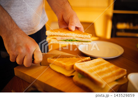Person slicing a sandwich with turkey and lettuce on a wooden cutting board, toasted bread and a white plate in a warm kitchen setting Person slicing a sandwich with turkey and lettuce on a wooden cutting board, toasted bread and a white plate in a warm kitchen setting 143529859