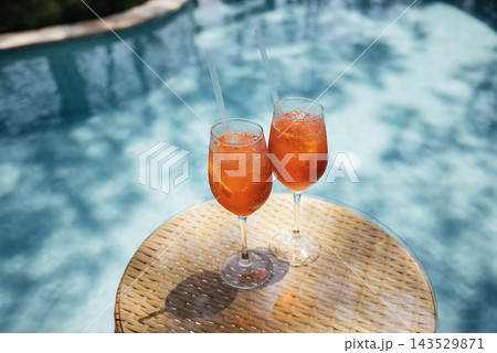 Two elegant glasses of orange cocktails with ice on a round wicker table beside a swimming pool, sunlight reflecting on the water surface in the background Two elegant glasses of orange cocktails with ice on a round wicker table beside a swimming pool, sunlight reflecting on the water surface in the background 143529871