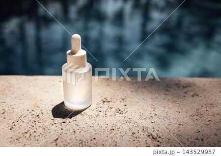 Clear glass dropper bottle with white cap placed on a textured stone surface beside a swimming pool, reflecting sunlight and creating soft shadows Clear glass dropper bottle with white cap placed on a textured stone surface beside a swimming pool, reflecting sunlight and creating soft shadows 143529987