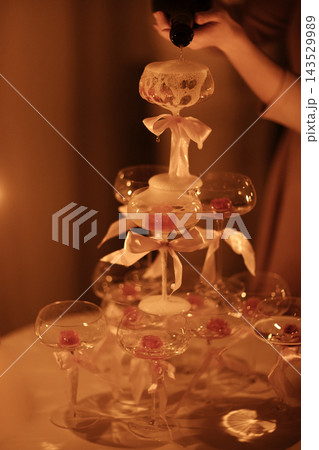 Champagne tower being filled with sparkling wine from a bottle, featuring elegant glass flutes adorned with pink ribbons, illuminated by soft ambient lighting Champagne tower being filled with sparkling wine from a bottle, featuring elegant glass flutes adorned with pink ribbons, illuminated by soft ambient lighting 143529989