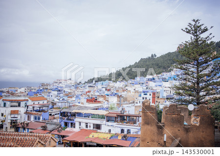 Blue-painted buildings and rooftops of Chefchaouen, Morocco, nestled against a mountainous backdrop under a cloudy sky, showcasing traditional architecture and vibrant colors Blue-painted buildings and rooftops of Chefchaouen, Morocco, nestled against a mountainous backdrop under a cloudy sky, showcasing traditional architecture and vibrant colors 143530001
