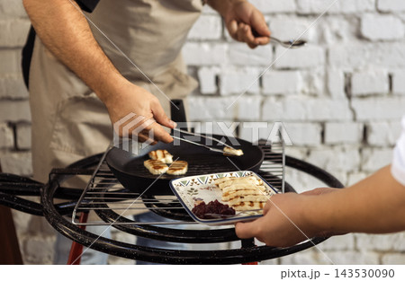 Cooking grilled cheese. Chef hands holding tongs and putting pieces of cheese on plate. Waiter holds tray with soft traditional Cypriot pickled haloumi and jam. Cooking grilled cheese. Chef hands holding tongs and putting pieces of cheese on plate. Waiter holds tray with soft traditional Cypriot pickled haloumi and jam. 143530090