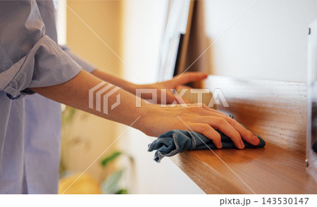 A young woman in a loose shirt wipes dust from a wooden shelf in living room 143530147