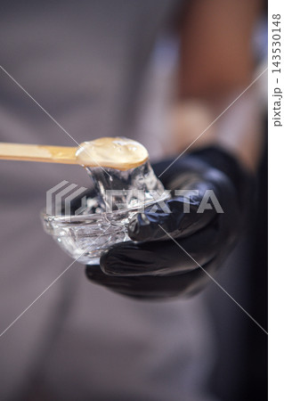 Close-up of a cosmetologist hands with a glass bowl and moisturizing gel. The dermatologist hands in black gloves hold a jar of nourishing serum and a wooden spatula. Close-up of a cosmetologist hands with a glass bowl and moisturizing gel. The dermatologist hands in black gloves hold a jar of nourishing serum and a wooden spatula. 143530148