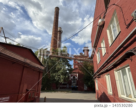 Historic red brick industrial courtyard with tall smokestacks, overhead pipes, blue sky, and clouds Historic red brick industrial courtyard with tall smokestacks, overhead pipes, blue sky, and clouds 143530280