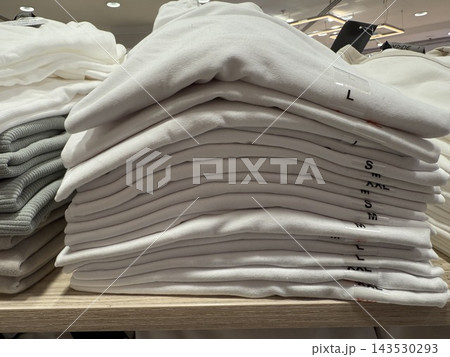 Stacked white folded clothing with visible size labels displayed on wooden retail shelving Stacked white folded clothing with visible size labels displayed on wooden retail shelving 143530293
