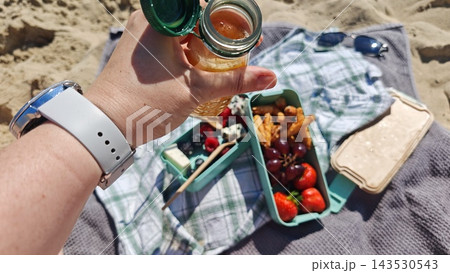 Hand holding an open glass jar of amber drink above a picnic spread with fruit, snacks, blanket, sunglasses, and sand at a sunny beach 143530543