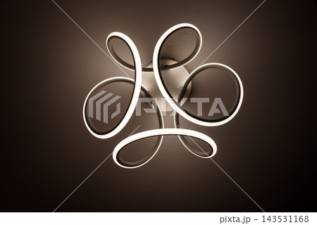 A modern LED ceiling light with looped rings illuminates a dark interior room. Minimal interior lighting, contemporary design, decoration, abstract shapes, architecture and technology. A modern LED ceiling light with looped rings illuminates a dark interior room. Minimal interior lighting, contemporary design, decoration, abstract shapes, architecture and technology. 143531168