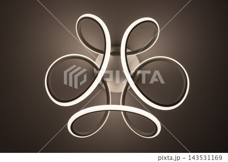 A modern LED ceiling light with looped rings illuminates a dark interior room. Minimal interior lighting, contemporary design, decoration, abstract shapes, architecture and technology. 143531169