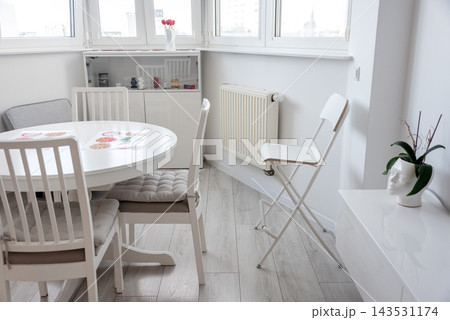 Bright minimalist dining and living area with white furniture and a potted plant by the window. Interior design, minimalism, home lifestyle, calm, simplicity, cozy morning. Bright minimalist dining and living area with white furniture and a potted plant by the window. Interior design, minimalism, home lifestyle, calm, simplicity, cozy morning. 143531174