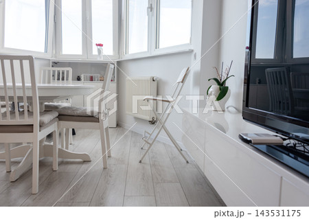 Bright minimalist dining and living area with white furniture and a potted plant by the window. Interior design, minimalism, home lifestyle, calm, simplicity, cozy morning. 143531175