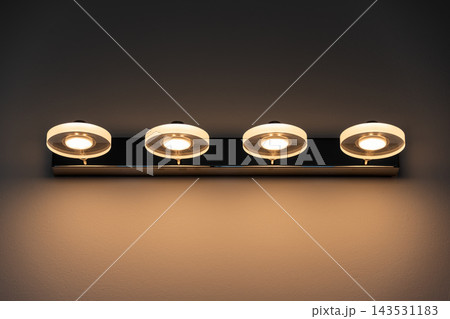 Four circular LED wall lights on a horizontal chrome bar illuminate a warm interior wall. Home lighting, modern design, minimalism, comfort, bathroom vanity, energy efficient, contemporary decor. 143531183