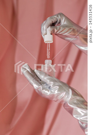 Hands in silver gloves holding a dropper above a clear bottle, with a soft pink fabric backdrop creating a modern and elegant aesthetic Hands in silver gloves holding a dropper above a clear bottle, with a soft pink fabric backdrop creating a modern and elegant aesthetic 143531416