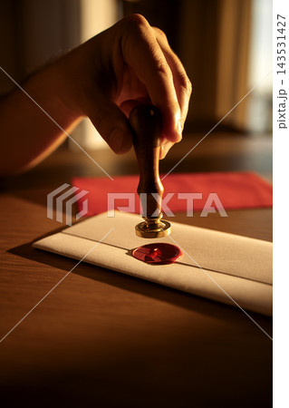 Hand applying a wax seal with a wooden stamp on a white envelope, red wax impression visible, with a blurred red envelope in the background 143531427