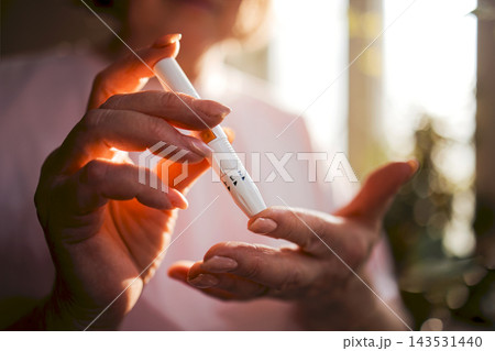 Elderly woman with light skin using a diabetes pen to check blood sugar levels, soft natural light illuminating her hands and the pen in a home setting 143531440