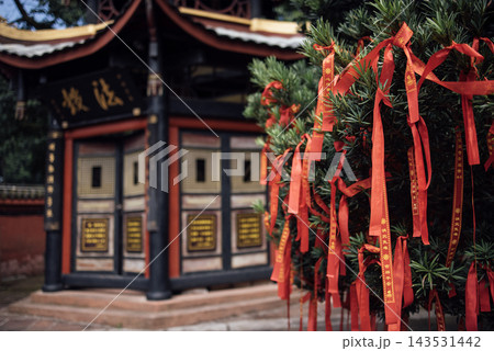 Red ribbons tied to a tree near a traditional Chinese temple with ornate architecture and gold inscriptions visible on the building's exterior Red ribbons tied to a tree near a traditional Chinese temple with ornate architecture and gold inscriptions visible on the building's exterior 143531442
