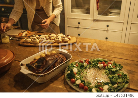 Woman preparing festive meal with roasted meat, decorated platter, and assorted cookies on wooden table in cozy kitchen setting Woman preparing festive meal with roasted meat, decorated platter, and assorted cookies on wooden table in cozy kitchen setting 143531486
