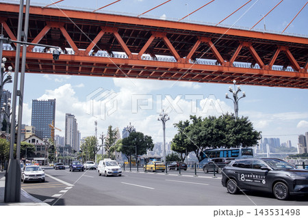 Urban cityscape featuring a busy street with vehicles, modern buildings, and a large orange bridge spanning over the road under a partly cloudy sky Urban cityscape featuring a busy street with vehicles, modern buildings, and a large orange bridge spanning over the road under a partly cloudy sky 143531498
