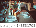 Elegant restaurant table setting with white plates, wine glasses, and a decorative centerpiece featuring greenery and flowers in a cozy dining environment 143531703
