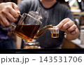 Person pouring tea from a glass teapot into a small cup, showcasing a warm beverage in a cozy indoor setting with blurred background elements 143531706