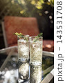 Two tall glasses of refreshing cocktail with ice and garnished with herbs placed on a glass table in a sunny outdoor setting with blurred background 143531708