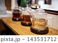 Three glass teapots filled with different types of tea displayed on a wooden countertop in a cozy tea shop setting with blurred background elements 143531712