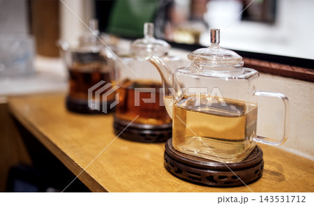 Three glass teapots filled with different types of tea displayed on a wooden countertop in a cozy tea shop setting with blurred background elements Three glass teapots filled with different types of tea displayed on a wooden countertop in a cozy tea shop setting with blurred background elements 143531712
