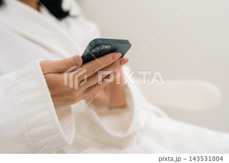 Close-up cropped shot of female patient in soft white bathrobe scrolling smartphone with manicured hands, using health and wellness apps while waiting for medical or spa appointment in modern clinic. 143531804