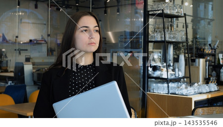 Young businesswoman holds closed modern laptop while standing inside bright modern coffee shop. Focused professional worker pauses with thoughtful expression during busy workday morning. Young businesswoman holds closed modern laptop while standing inside bright modern coffee shop. Focused professional worker pauses with thoughtful expression during busy workday morning. 143535546