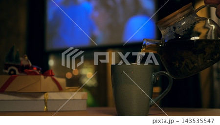 Aromatic beverage flows into ceramic cup from glass pot during cozy holiday evening indoors. Holiday presents with decorative ribbons rest nearby against backdrop of glowing screen at night. 143535547