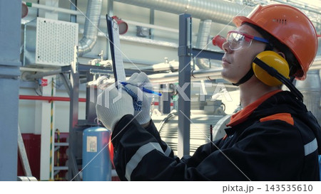 Focused technician in hard hat records inspection notes on clipboard inside industrial facility. Technician wearing gloves, goggles, and hearing protection stands near pipes and machinery. 143535610