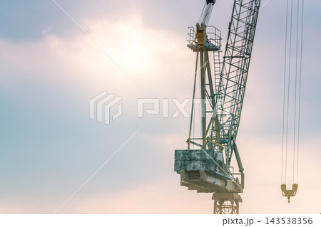Construction tower crane with a lattice boom, suspended hook and concrete counterweights against a blue and pink sky, heavy lifting equipment for building projects and real estate development. Construction tower crane with a lattice boom, suspended hook and concrete counterweights against a blue and pink sky, heavy lifting equipment for building projects and real estate development. 143538356