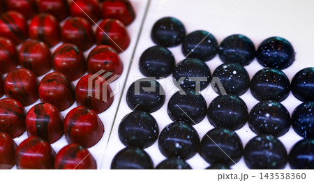 Dark chocolate pralines with raspberry and cognac ganache fillings on white plates, handcrafted bonbons with red shells and dark rounded shells decorated with speckles, a luxury dessert assortment. Dark chocolate pralines with raspberry and cognac ganache fillings on white plates, handcrafted bonbons with red shells and dark rounded shells decorated with speckles, a luxury dessert assortment. 143538360