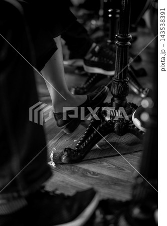 Foot wearing a leather loafer resting against an ornate metal pedestal base on a wooden floor, with other people wearing sneakers nearby and floral decoration around the fluted support column. Foot wearing a leather loafer resting against an ornate metal pedestal base on a wooden floor, with other people wearing sneakers nearby and floral decoration around the fluted support column. 143538391