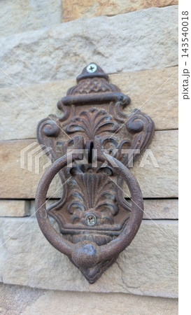 Ornate rusted metal door knocker mounted on textured stone wall, close view of vintage decorative hardware with weathered circular ring, aged finish, and carved floral detail Ornate rusted metal door knocker mounted on textured stone wall, close view of vintage decorative hardware with weathered circular ring, aged finish, and carved floral detail 143540018