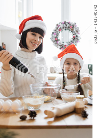 A mother and daughter wearing Santa hats joyfully bake Christmas cookies together in a bright kitchen A mother and daughter wearing Santa hats joyfully bake Christmas cookies together in a bright kitchen 143540242