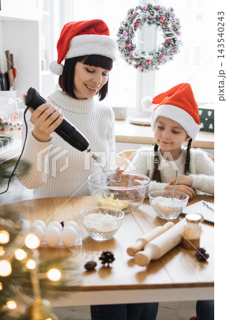 A mother and daughter wearing Santa hats bake together in a festive kitchen preparing holiday treats 143540243