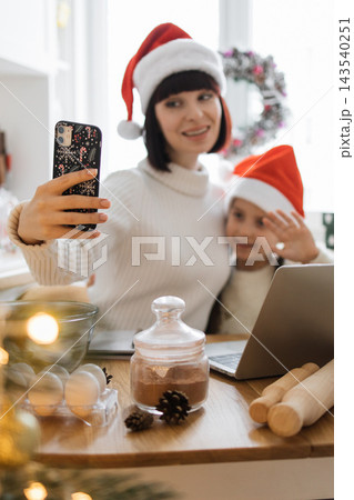 A mother and child wearing Santa hats take a selfie on a smartphone while preparing Christmas treats in the kitchen A mother and child wearing Santa hats take a selfie on a smartphone while preparing Christmas treats in the kitchen 143540251