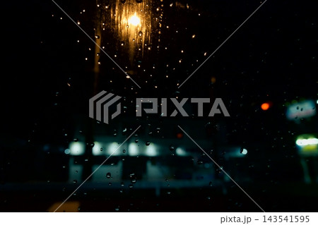 雨粒が付いた窓ガラス越しの夜の街灯と、ぼやけた店舗の明かり 雨粒が付いた窓ガラス越しの夜の街灯と、ぼやけた店舗の明かり 143541595
