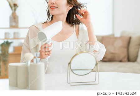 家のリビングでドライヤーを使って髪を乾かす女性。お風呂上がりのヘアケアと美容習慣 家のリビングでドライヤーを使って髪を乾かす女性。お風呂上がりのヘアケアと美容習慣 143542624