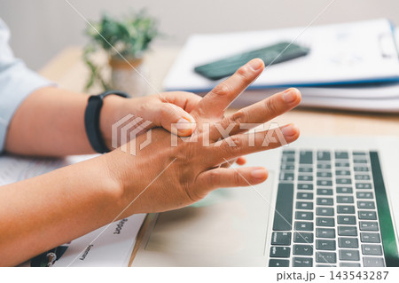 Office worker massaging the back of hand and fingers beside laptop after prolonged typing, illustrating repetitive strain, hand fatigue, workplace ergonomics, overuse discomfort 143543287